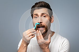 Handsome middle aged bearded man shaving mustache with razor and looking at camera over grey background