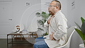 Handsome middle-aged caucasian man, with beard and glasses, sitting unhappily in chair, worriedly coughing in waiting room amid