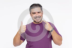 A handsome middle aged man pointing to his teeth and smiling while making a thumbs up sign. Dental concept. Wearing a purple
