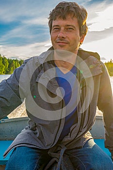 Handsome middle-aged man sitting at boat stern and floating along northern river on beautiful landscape background at sunset.