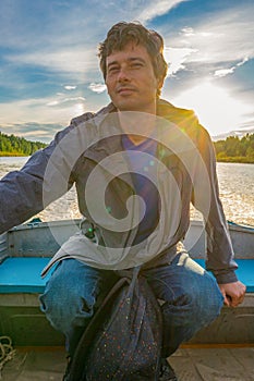 Handsome middle-aged man sitting at boat stern and floating along northern river on beautiful landscape background at sunset.