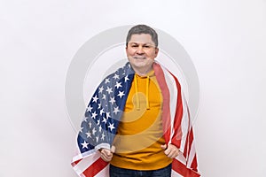 Handsome middle aged man standing wrapped in USA flag, celebrating national holiday.