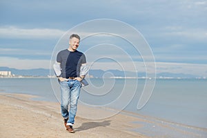Handsome middle-aged man walking at the beach. Attractive happy smiling mid adult male model posing at seaside in blue jeans, t-