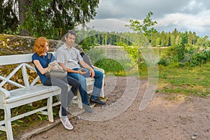 Handsome middle-aged man and young pretty lady sitting on bench and using smartphones in summer evening. Tourists on the beautiful