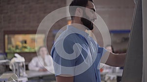 Handsome Middle Eastern man with black hair and beard writing on board and greeting students in dental audience. Smart