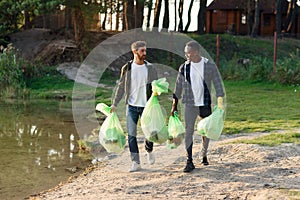 Handsome mixed race active friends carrying plastic bags near the lake after cleanup surrounding territory from rubbish.