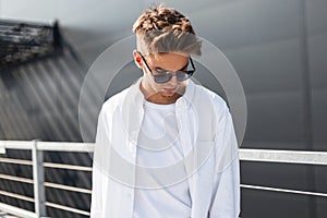 Handsome modern young man in fashionable white clothes in stylish black sunglasses with trendy hairstyle stands on a bright sunny
