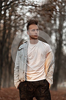 Handsome modern young man in a vintage white T-shirt in a blue denim jacket with a fashionable hairstyle posing outdoors in a park