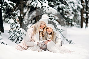 Handsome mother and daughter are having fun outdoor in winter time. Sitting on snow in forest between snowy trees