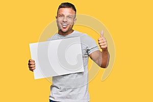 Handsome muscle man holding blank empty banner smiling happy and positive, thumb up doing excellent and approval sign