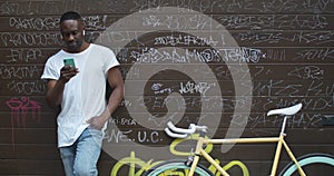 Handsome Muscular African American Young Guy in Earphones Using his Smartphone while Standing near Stylish Modern Bike
