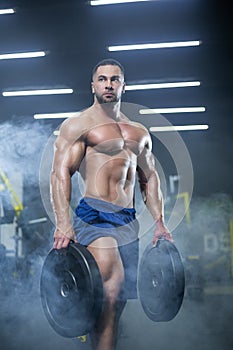 Handsome muscular athlete man is standing in a gym against the background of smoke holding barbell discs in both hands