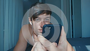 Handsome naked smiling man with headphones lying in bed at home and scrolling on smartphone