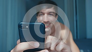 Handsome naked smiling man lying in bed at home and using smartphone