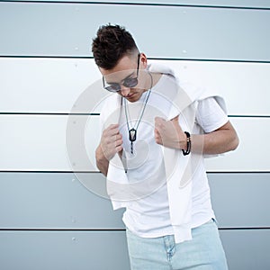 Handsome nice young hipster man in trendy white t-shirt in stylish sunglasses with fashionable hairstyle posing near a modern