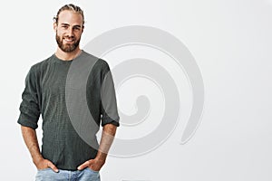 Handsome nordic man with beard and stylish hairstyle in grey shirt and jeans smiling, looking in camera, keeps hands in