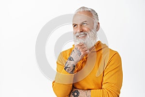 Handsome old man with tattoos, touch his beard thoughtful, smiling and looking satisfied at upper left corner empty