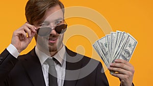 Handsome oligarch in sunglasses showing dollars and winking, wealth, close-up