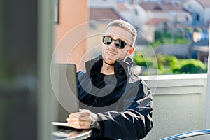 Handsome pensive stylish man sitting on balcony and working on laptop
