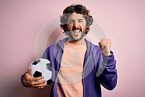 Handsome player man with beard playing soccer holding football ball over pink background screaming proud and celebrating victory