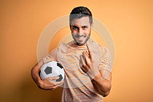 Handsome player man with beard playing soccer holding footballl ball over yellow background Beckoning come here gesture with hand