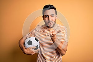 Handsome player man with beard playing soccer holding footballl ball over yellow background looking at the camera blowing a kiss