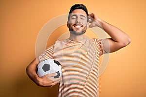 Handsome player man with beard playing soccer holding footballl ball over yellow background smiling confident touching hair with