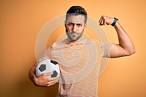 Handsome player man with beard playing soccer holding footballl ball over yellow background Strong person showing arm muscle,