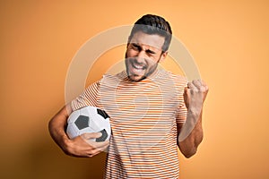 Handsome player man with beard playing soccer holding footballl ball over yellow background very happy and excited doing winner