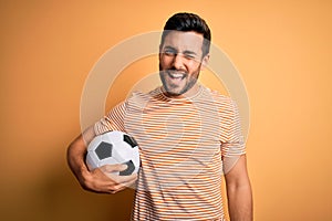 Handsome player man with beard playing soccer holding footballl ball over yellow background winking looking at the camera with