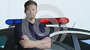 Handsome policeman standing near cop car and looking into camera, on duty