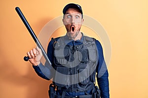 Handsome policeman wearing uniform and bulletprof holding baton over yellow background scared and amazed with open mouth for