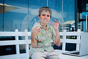 Handsome pre teen boy in summer shirt, holding a glass of freshy squeezed orange juice, showing OK hand sign at camera