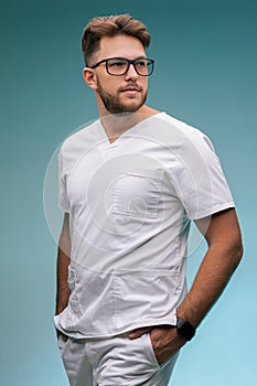 Handsome professional young male physician practitioner in glasses and medical uniform on blue studio background