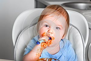 Handsome red-haired baby eating in children`s chair. First feeding children with porridge, vegetables, fruits, meat