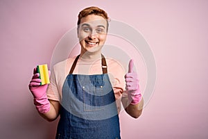 Handsome redhead man doing housework wearing apron and gloves using cleaner scourer Smiling happy and positive, thumb up doing