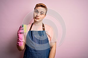Handsome redhead man doing housework wearing apron and gloves using cleaner scourer thinking attitude and sober expression looking