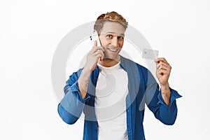 Handsome redhead man making phone call to bank support, showing credit card and smiling carefree, using contactless