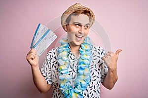 Handsome redhead tourist man on vacation wearing hawaiian lei holding boarding pass airlane pointing thumb up to the side smiling