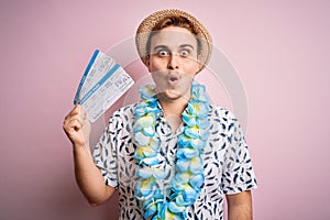 Handsome redhead tourist man on vacation wearing hawaiian lei holding boarding pass airlane scared and amazed with open mouth for