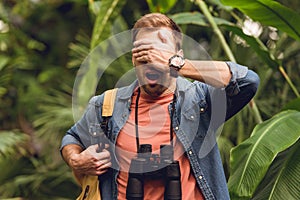 Handsome scared traveler with binoculars and backpack putting hand on eyes in green tropical forest.