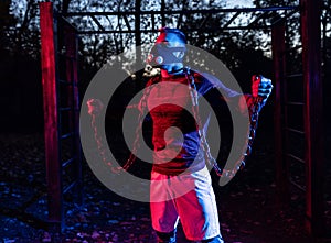 Handsome scary muscular man working out with weightlifting chains at dusk in the park while wearing training mask, functional