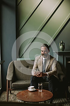 Handsome senior businessman drinking coffee and using mobile phone in lobby