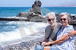Handsome senior couple sitting on the rocks looking at camera smiling. Two retired enjoying summer vacation and freedom