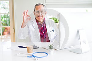 Handsome senior doctor man doing research using laptop looking for a cure at the clinic doing ok sign with fingers, excellent