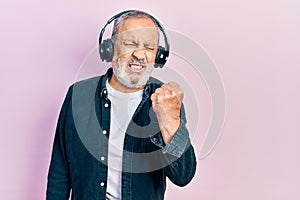 Handsome senior man with beard listening to music using headphones angry and mad raising fist frustrated and furious while