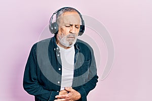 Handsome senior man with beard listening to music using headphones with hand on stomach because indigestion, painful illness