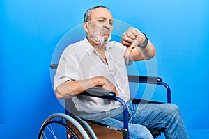 Handsome senior man with beard sitting on wheelchair looking unhappy and angry showing rejection and negative with thumbs down