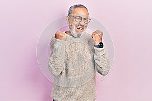 Handsome senior man with beard wearing casual sweater and glasses angry and mad raising fists frustrated and furious while