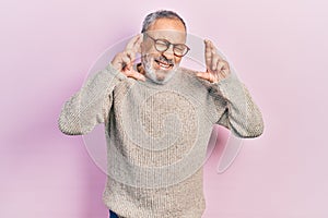 Handsome senior man with beard wearing casual sweater and glasses gesturing finger crossed smiling with hope and eyes closed